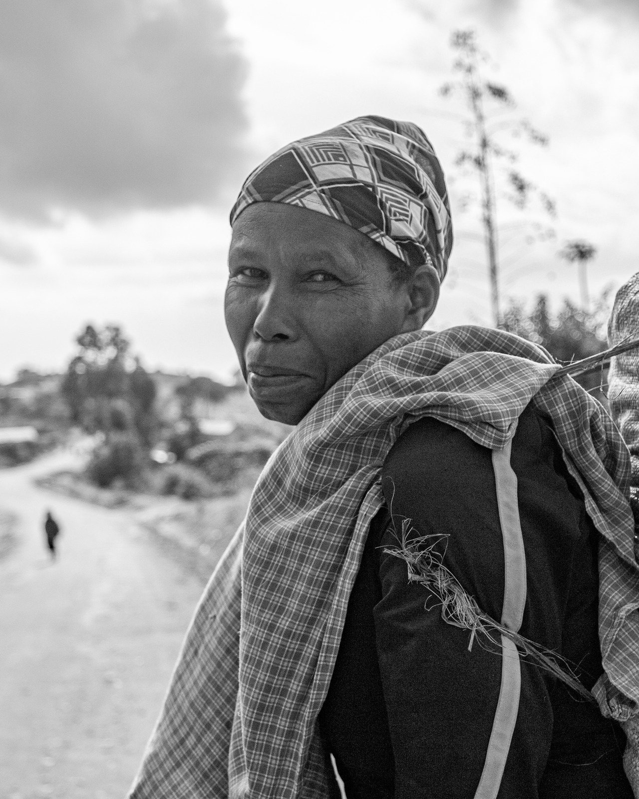 Black and white portrait of a woman in a patterned headscarf looking over her shoulder on a rural road